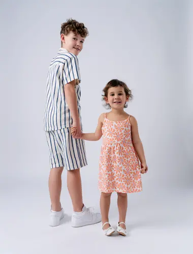 Ein Junge in einem gestreiften Outfit und ein Mädchen in einem geblümten Kleid halten sich lächelnd an den Händen, vor einem schlichten weißen Hintergrund.