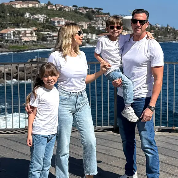 Eine vierköpfige Familie posiert gemeinsam am Meer, trägt passende weiße T-Shirts und Jeans, mit einer malerischen Kulisse aus Hügeln und Wasser im Hintergrund.