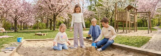 Vier Kinder spielen und bauen Sandburgen in einem Sandkasten unter blühenden Bäumen in einem Park.
