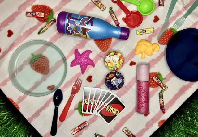 Ein buntes Picknick mit Tellern, Utensilien, Süßigkeiten, Spielzeug und einer Trinkflasche auf einer Decke mit Erdbeermuster.