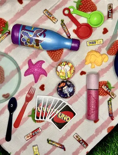 Ein buntes Picknick mit Tellern, Utensilien, Süßigkeiten, Spielzeug und einer Trinkflasche auf einer Decke mit Erdbeermuster.