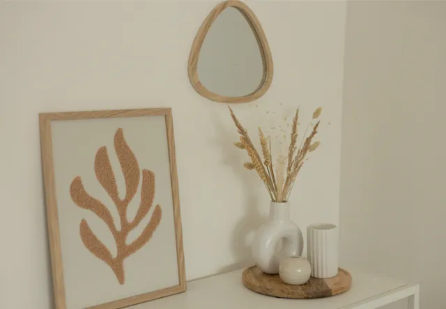 Eine minimalistische Einrichtung mit einem kleinen Spiegel, einem gerahmten Blattkunstwerk und einer Vase mit Trockenblumen auf einem Holztablett vor einer neutralen Wand.