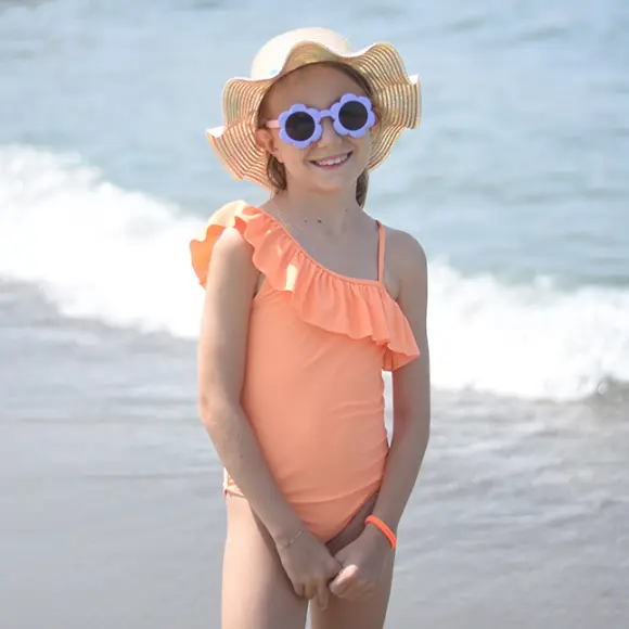 Ein Kind im korallfarbenen One-Shoulder-Badeanzug mit Rüschen steht am Strand und trägt einen breitkrempigen Strohhut