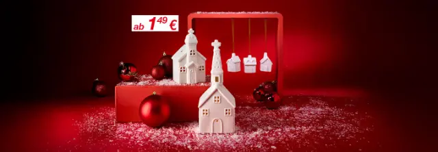 Weiße Keramikhäuser und Ornamente vor rotem Hintergrund, bestreut mit schneeähnlichen Verzierungen, schaffen eine festliche Weihnachtsstimmung.