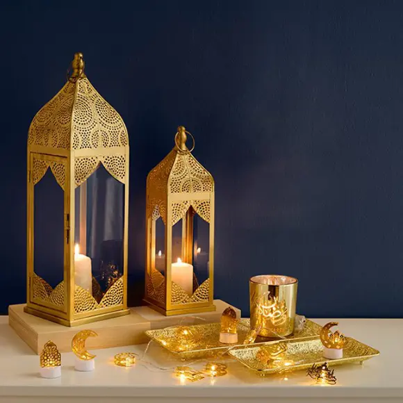 Zwei aufwendig gestaltete goldene Laternen mit Kerzen, ein Ziertablett und festliche Lichter vor einer tiefblauen Wand.