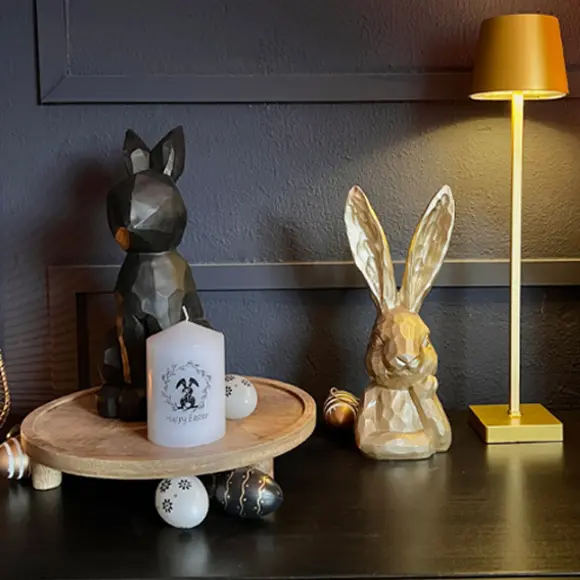 Ein österliches Holz-Arrangement mit zwei Hasendekorationen, einer Kerze und Dekoeiern auf einem runden Tablett vor dunklem Hintergrund.