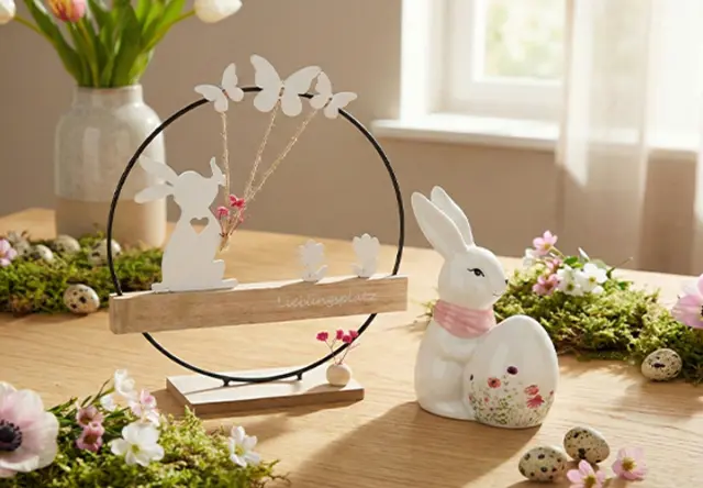 Eine dekorative Tischdekoration mit einer weißen Hasenfigur, einem Holzdisplay mit Schmetterlingen und floralen Akzenten auf Grünpflanzen.