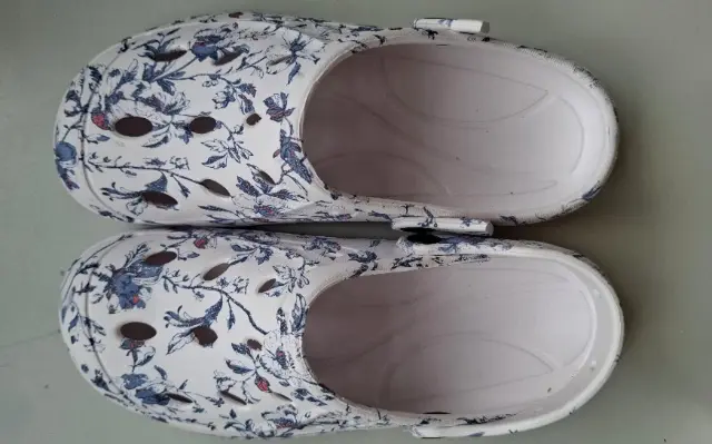 Weiße Clogs mit Blumenmuster und Belüftungslöchern, von oben gesehen, auf einer ebenen Fläche stehend.