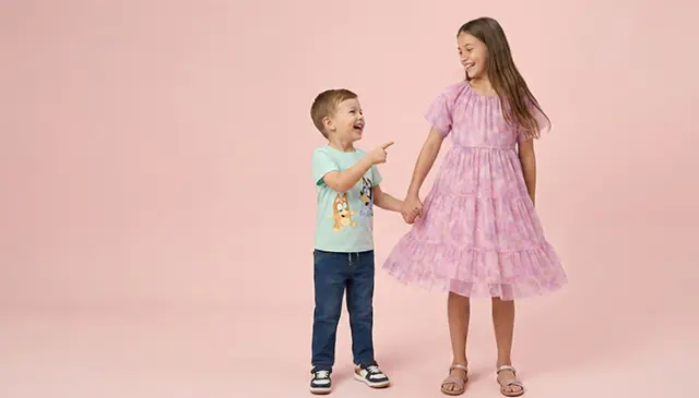 Ein kleiner Junge in einem hellblauen Hemd und Jeans zeigt spielerisch auf ein Mädchen in einem rosa Rüschenkleid, beide vor einem zartrosa Hintergrund. Ein kleiner Junge in einem hellblauen Hemd und Jeans zeigt spielerisch auf ein Mädchen in einem rosa Rüschenkleid, beide vor einem zartrosa Hintergrund.