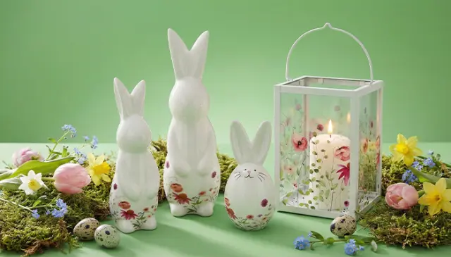 Drei weiße Keramikhasen, umgeben von bunten Blumen, Eiern und einer Blumenlaterne mit einer Kerze, vor einem hellgrünen Hintergrund. Drei weiße Keramikhasen, umgeben von bunten Blumen, Eiern und einer Blumenlaterne mit einer Kerze, vor einem hellgrünen Hintergrund.