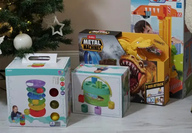 Eine Sammlung bunter Kinderspielzeuge: Stapelringe, ein Spielset und ein Dinosaurier-Spiel, arrangiert neben einem geschmückten Weihnachtsbaum.
