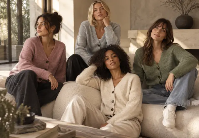 Herbstliche Strick-Outfits für Damen Vier Frauen tragen Strickjacken in Rosa, Grau, Creme und Grün in einer gemütlichen Wohnzimmerszene.