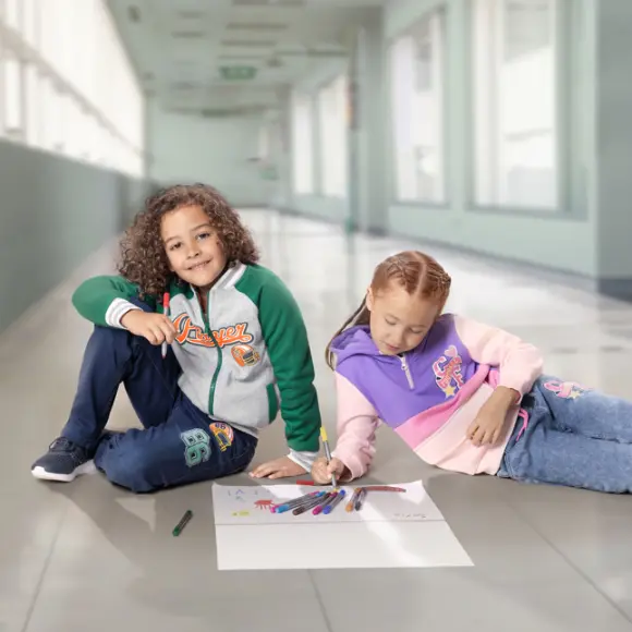 Zwei Kinder sitzen auf einem gefliesten Boden und malen mit farbigen Stiften auf einem großen Blatt Papier.