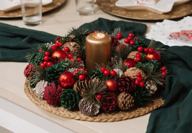 Ein festlicher Weihnachtsmittelpunkt mit einer goldenen Kerze, umgeben von Tannenzapfen, roten Beeren und Christbaumschmuck auf einem geflochtenen Untersetzer. Ein festlicher Weihnachtsmittelpunkt mit einer goldenen Kerze, umgeben von Tannenzapfen, roten Beeren und Christbaumschmuck auf einem geflochtenen Untersetzer.