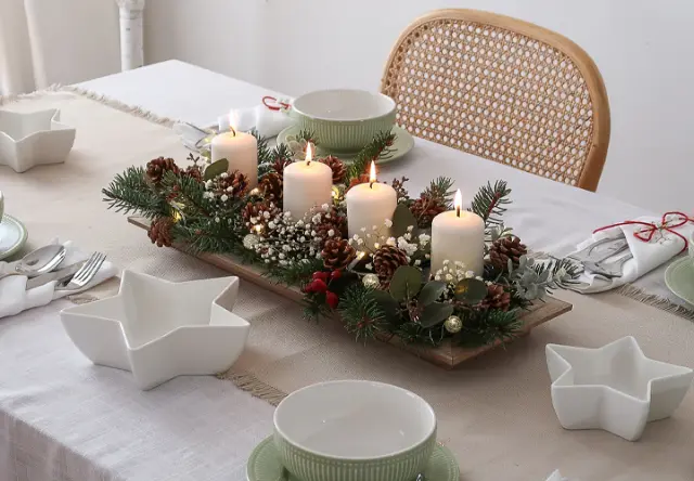 Ein festlich gedeckter Tisch mit einem Mittelstück aus brennenden Kerzen, umgeben von Grün, Tannenzapfen und weißen Blumen auf einer neutralen Tischdecke. Ein festlich gedeckter Tisch mit einem Mittelstück aus brennenden Kerzen, umgeben von Grün, Tannenzapfen und weißen Blumen auf einer neutralen Tischdecke.