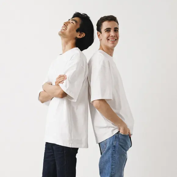 Basic Herrenmode Zwei junge Männer in weißen T-Shirts stehen Rücken an Rücken vor einem schlichten weißen Hintergrund und lächeln entspannt.