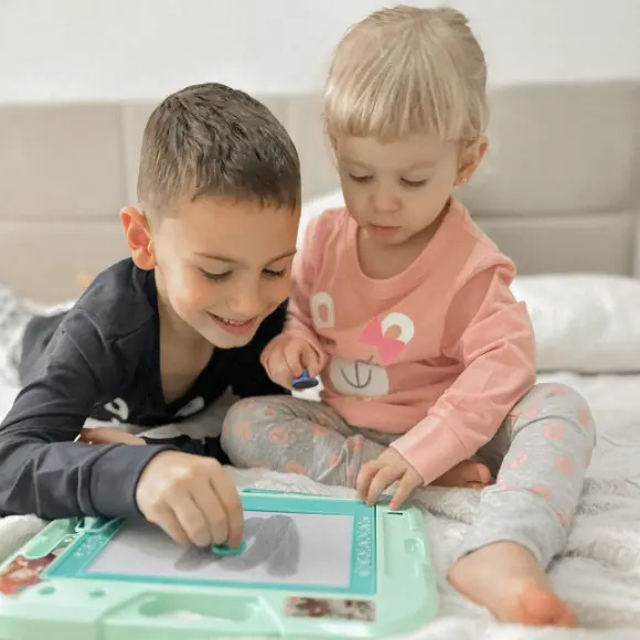 Zwei kleine Kinder sitzen auf einem Bett, spielen fröhlich miteinander und mit einem Zeichentablet, umgeben von weicher Bettwäsche.