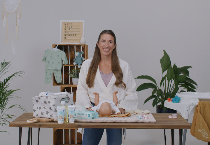 Eine Frau steht an einem Holztisch mit Babypflegeprodukten, Kleidung und Spielzeug, umgeben von Grünpflanzen in einer hellen Umgebung.