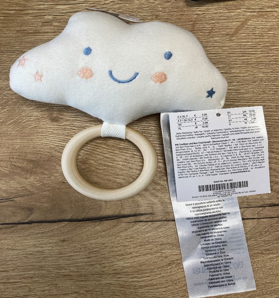 Ein weiches, plüschiges Babyspielzeug in Wolkenform mit lächelndem Gesicht, Holzring und angenähtem Pflegeetikett auf einer Holzoberfläche.