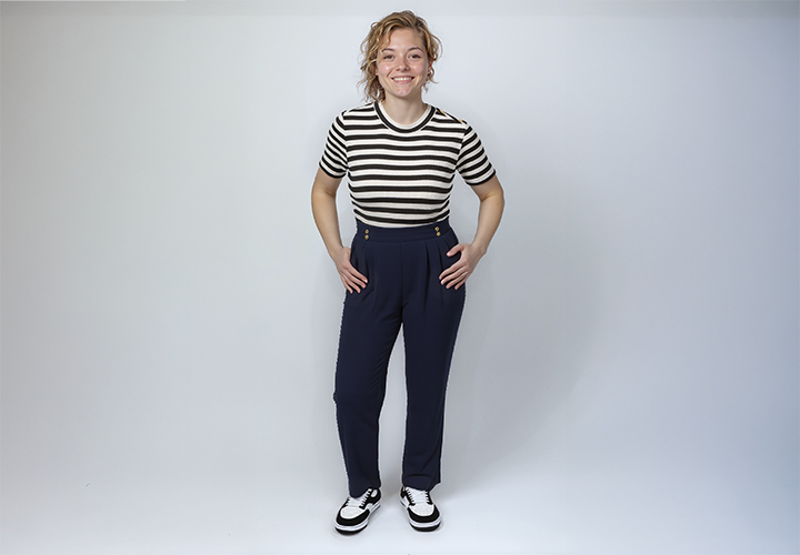 Eine Person steht selbstbewusst in einem gestreiften Shirt und einer hoch taillierten marineblauen Hose, kombiniert mit schwarz-weißen Schuhen, vor einem schlichten Hintergrund.