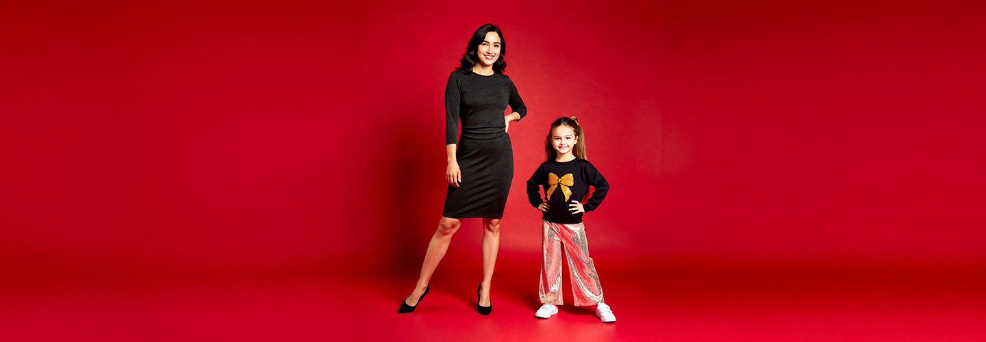 Eine Frau in einem eng anliegenden schwarzen Kleid steht neben einem Mädchen in einem schwarzen Hemd und glänzenden silbernen Hosen, beide vor einem leuchtend roten Hintergrund.