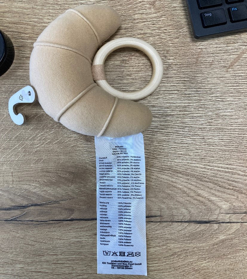 Ein weiches, croissantförmiges Plüschtier mit Holzring und Pflegeetikett, auf einer Holzoberfläche neben einer Tastatur liegend.