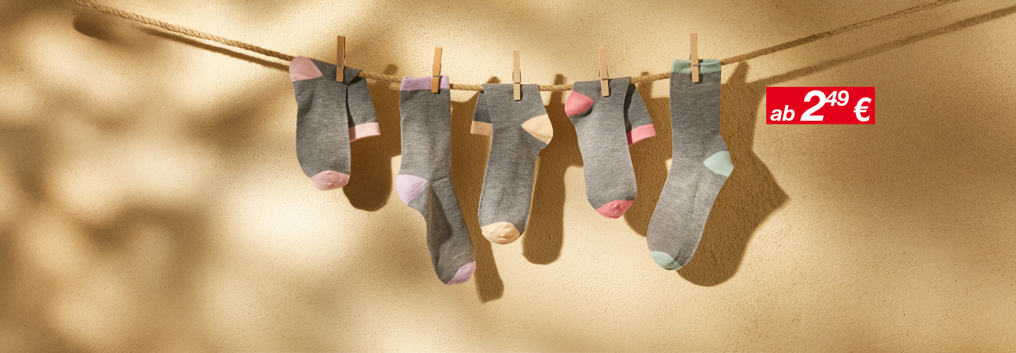Fünf Paar graue Socken mit pastellfarbenen Zehen hängen an einer Wäscheleine vor einer sanft gelben Wand, beleuchtet von warmem Licht.