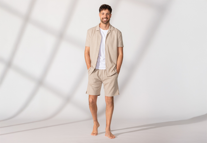 Ein barfuß stehender Mann trägt ein beigefarbenes Kurzarmhemd mit Knöpfen und passende Shorts, die Hände lässig in den Taschen – vor hellem Hintergrund.