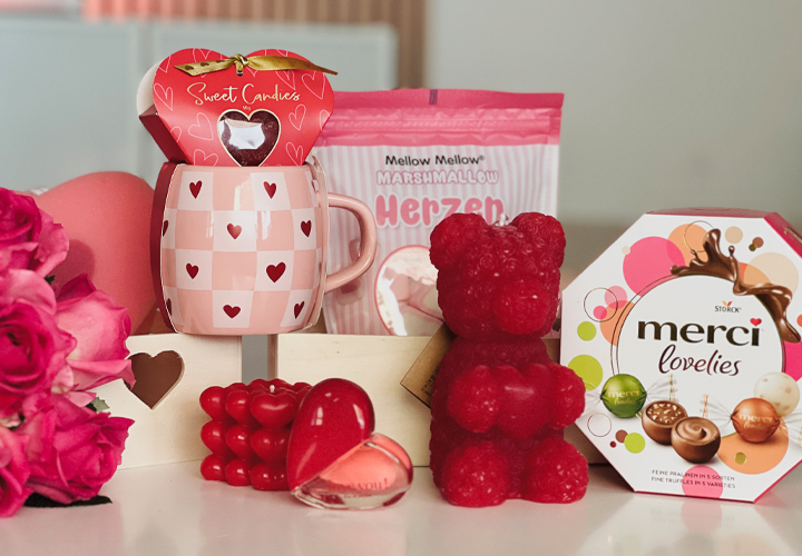 Zeig deine Liebe! Eine liebevoll gestaltete Valentinstagsauslage mit einer Tasse im Herzdesign, Süßigkeiten, einem roten Gummibär und einer Schachtel Pralinen – umrahmt von Rosen.