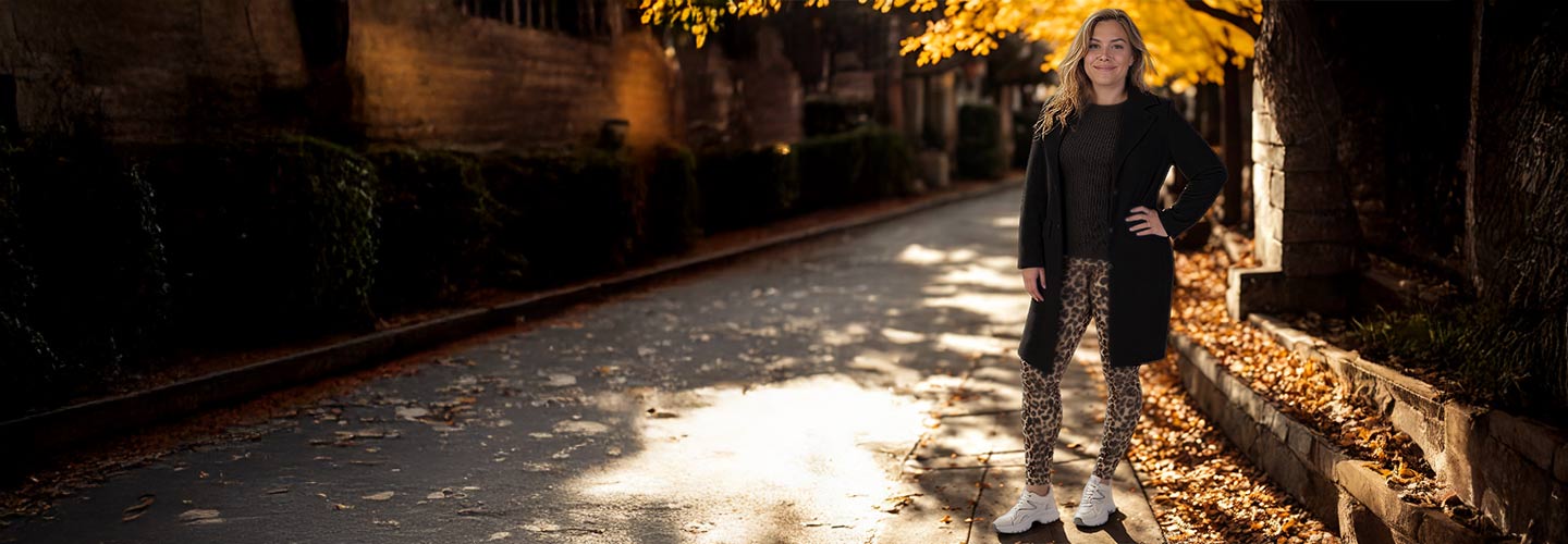 Eine Person steht selbstbewusst auf einer mit Laub bedeckten Straße, trägt einen dunklen Mantel, gemusterte Leggings und weiße Turnschuhe und ist von Herbstlaub umgeben.