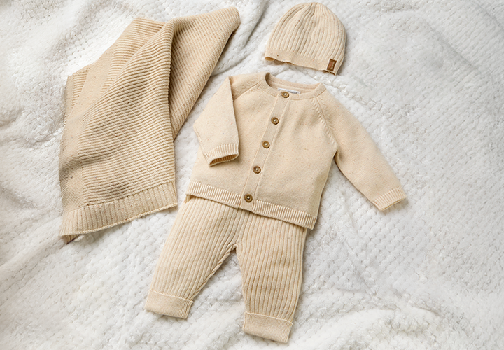 Ein weiches Babyoutfit in Beige mit Strickjacke, Hose und passender Mütze, präsentiert auf einer strukturierten Decke.