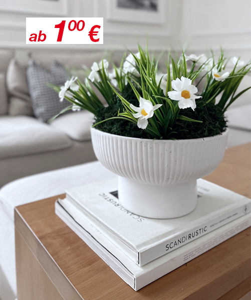 Eine weiße Schale mit künstlichen Gänseblümchen und Grün steht auf gestapelten Büchern auf einem Holzcouchtisch, im Hintergrund ein helles Sofa.